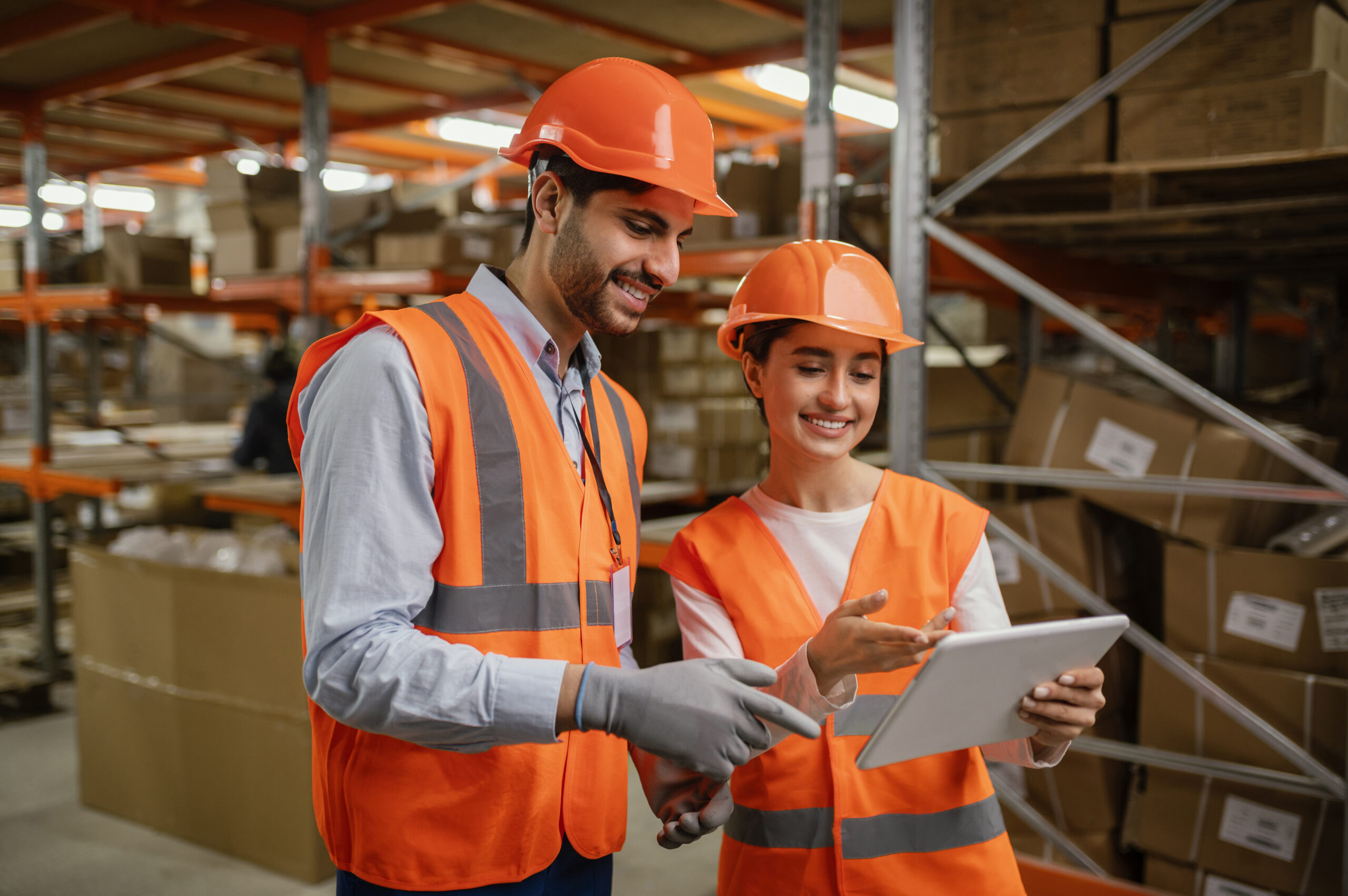 Two warehouse workers in orange vests and hard hats looking at a tablet in a storage facility.