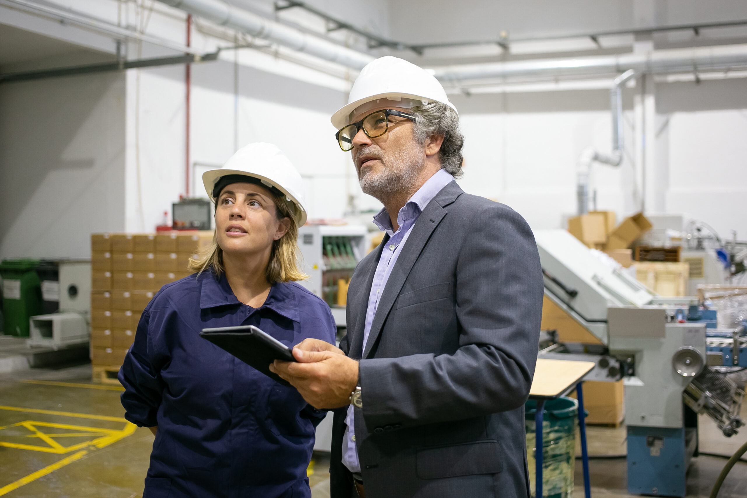 Two people, one woman and one man, wearing hard hats, looking up and holding a tablet in an industrial setting.