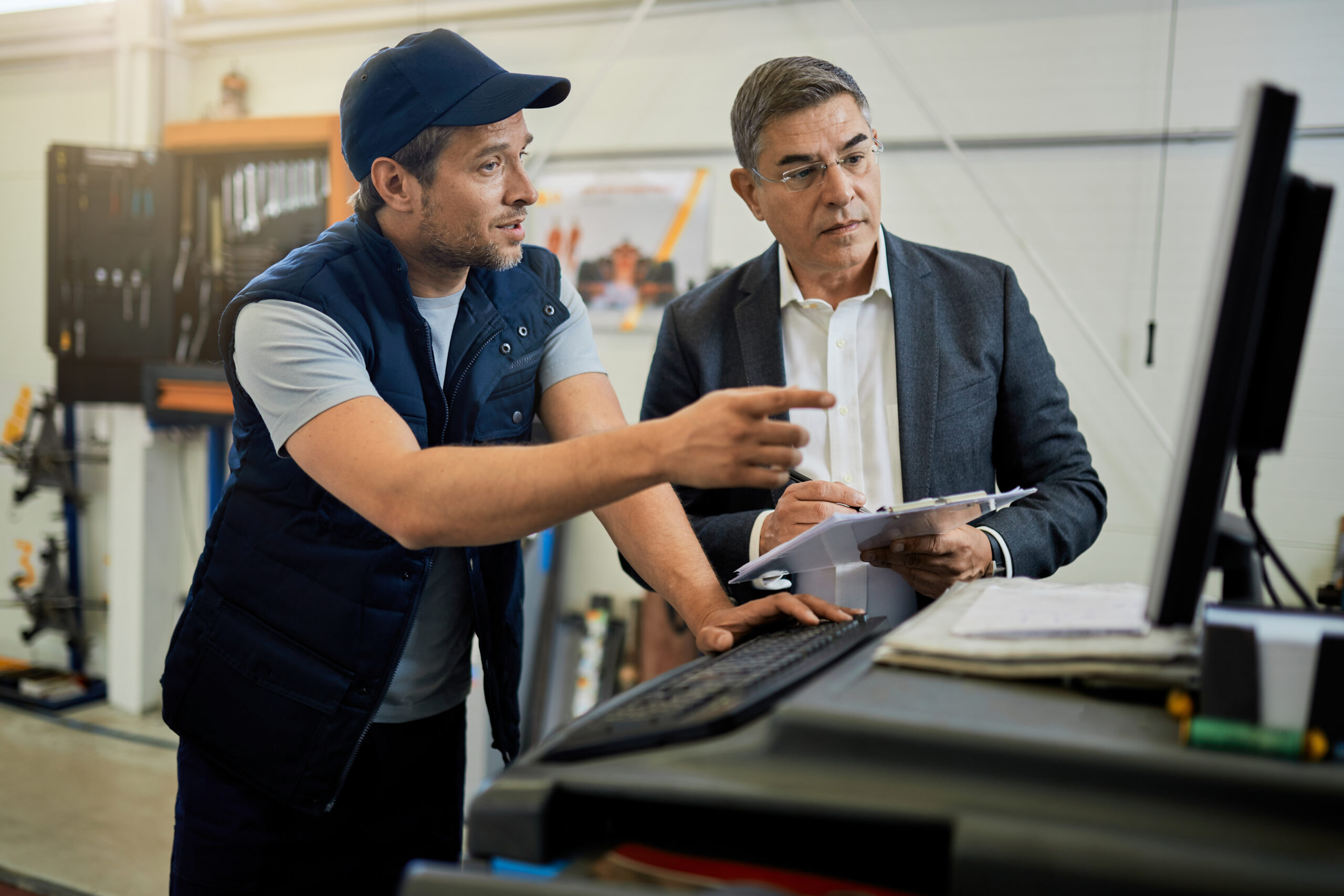 Two men, one in a blue cap and vest and the other in a suit, looking at a computer screen in a workshop or office setting.